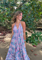 A woman standing in a natural setting wearing a long sleeveless dress with a floral pattern in blue and pink shades. The dress has an open back and is tied with strings around the neck and back.
